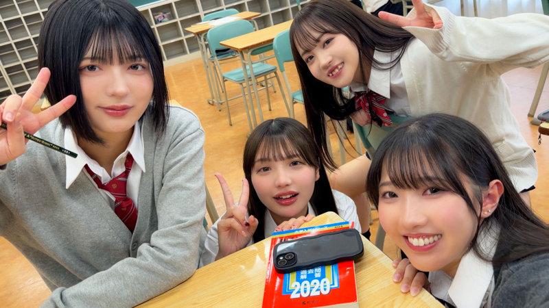 制服姿の4人組が教室の机を囲み、参考書とスマホの前でカメラを見る